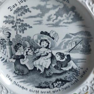 May include: White ceramic plate with a black and white illustration of children playing outdoors. The plate has decorative floral and scrollwork around the rim, and German text. A charming vintage piece.