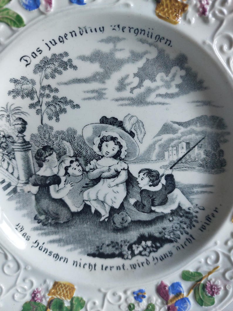 May include: White ceramic plate with a black and white illustration of children playing outdoors. The plate has decorative floral and scrollwork around the rim, and German text. A charming vintage piece.