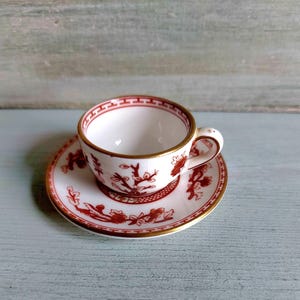Puede incluir: Un juego de taza y platillo pequeños, blancos con un diseño floral rojo y ribetes dorados. El conjunto se encuentra sobre una superficie azul claro. La taza tiene un asa pequeña.