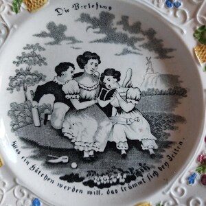 May include: White ceramic plate with a black and white pastoral scene featuring three figures and a windmill. The plate has a decorative floral border with blue, yellow, and pink accents, and German text.