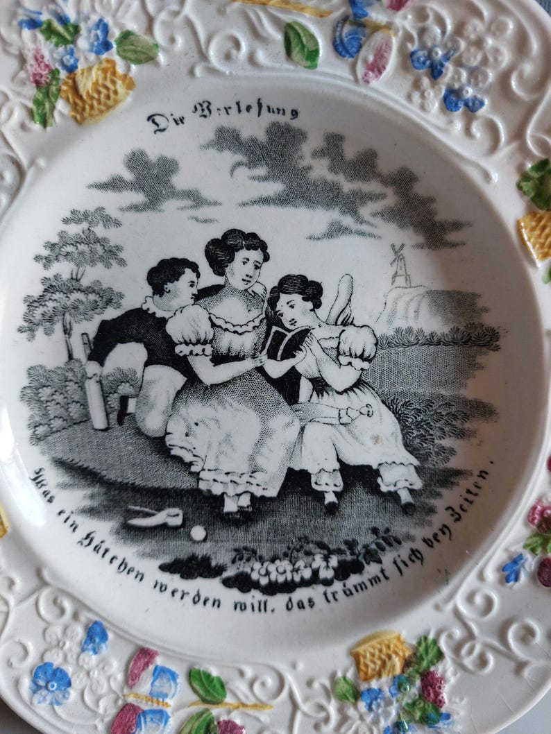May include: White ceramic plate with a black and white pastoral scene featuring three figures and a windmill. The plate has a decorative floral border with blue, yellow, and pink accents, and German text.