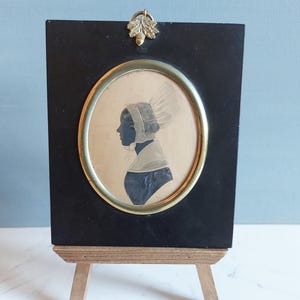 May include: A framed silhouette portrait of a woman in profile, set within an oval frame with a gold-coloured border. The portrait is mounted on a black rectangular base, supported by a small wooden easel. A decorative gold-coloured acorn and leaf adorn the top.