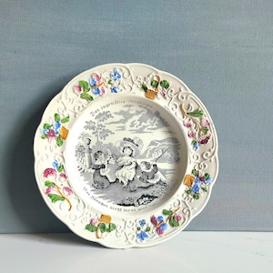 May include: White decorative plate with a scalloped edge, featuring a black and white pastoral scene and floral accents in pink, blue, and yellow. The plate has German text around the rim.