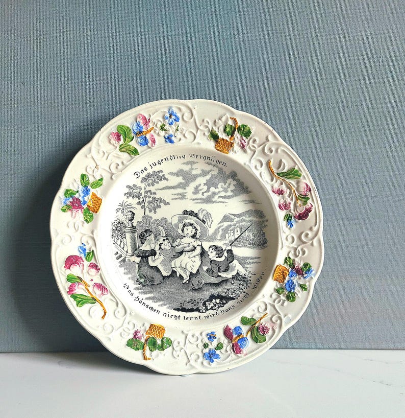May include: White decorative plate with a scalloped edge, featuring a black and white pastoral scene and floral accents in pink, blue, and yellow. The plate has German text around the rim.