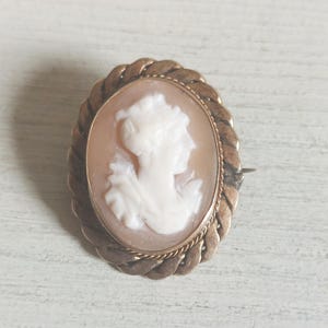 May include: An oval-shaped brooch featuring a carved white shell cameo of a profile portrait set against a light brown background. The cameo is framed by a twisted gold-toned metal border with a rope-like texture and a thin pin mechanism on the back.