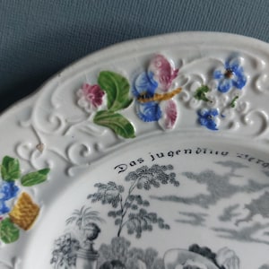 May include: White ceramic plate with embossed floral and butterfly details. The plate features a black and white pastoral scene and the text "Das jugendliche Herz". Decorative tableware.