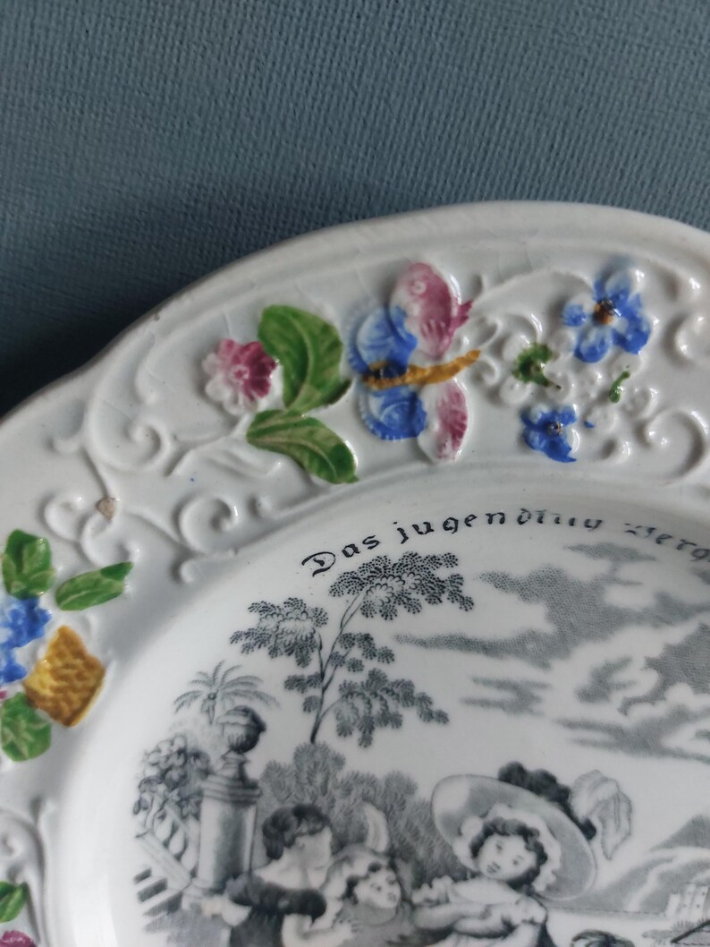 May include: White ceramic plate with embossed floral and butterfly details. The plate features a black and white pastoral scene and the text "Das jugendliche Herz". Decorative tableware.