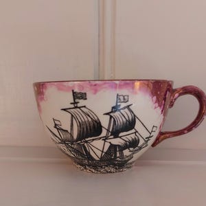 Könnte beinhalten: Eine weiße Teetasse mit einem weinroten Rand und Henkel, mit einer schwarz-weißen Illustration eines Segelschiffs. Die Tasse hat eine breite, abgerundete Form und steht auf einer weißen Oberfläche.
