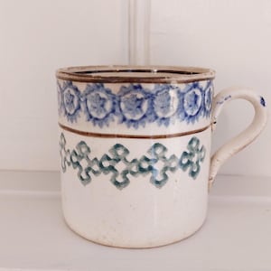 Pode incluir: Uma caneca de cerâmica branca com padrões florais azuis e verdes. A caneca tem uma alça curva e uma borda marrom. O design apresenta um padrão repetitivo de flores azuis e formas geométricas verdes.