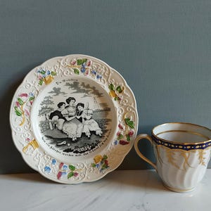 May include: A vintage porcelain plate and teacup set. The plate features a black and white illustration of people and text, with floral and scrollwork trim. The teacup is white with gold accents and a blue band.