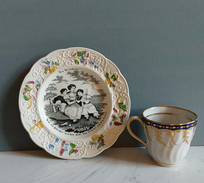 May include: A vintage porcelain plate and teacup set. The plate features a black and white illustration of people and text, with floral and scrollwork trim. The teacup is white with gold accents and a blue band.