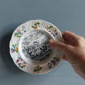 May include: A white decorative plate with a scalloped edge, featuring a black and white pastoral scene and floral accents in pink, blue, and yellow. The plate has text around the rim.