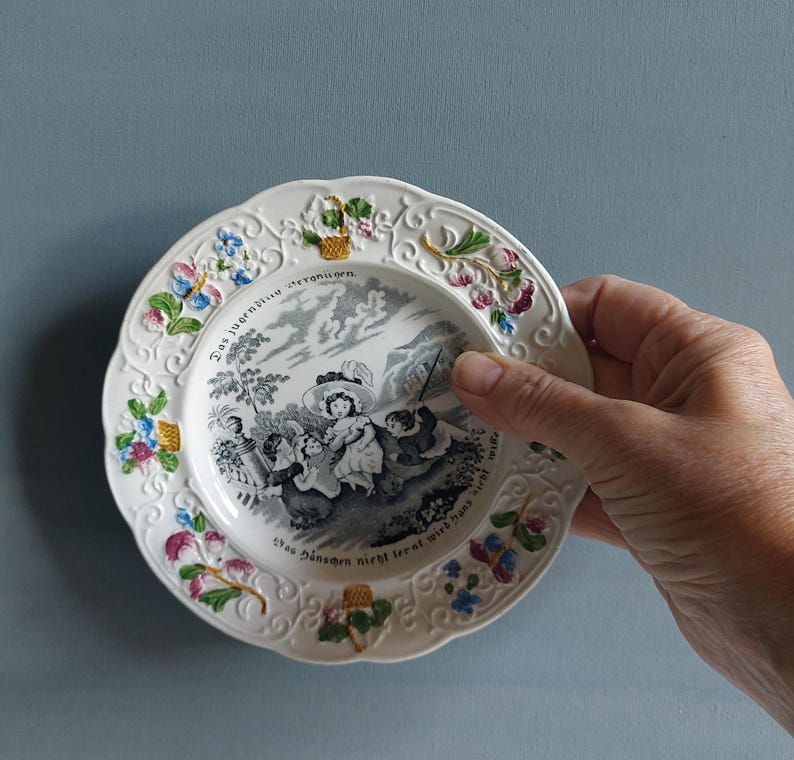 May include: A white decorative plate with a scalloped edge, featuring a black and white pastoral scene and floral accents in pink, blue, and yellow. The plate has text around the rim.