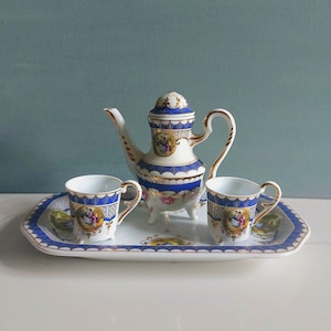 Peut inclure: Service à thé en porcelaine vintage comprenant une théière, deux tasses et un plateau. L'ensemble présente une base blanche avec des bordures bleues, des accents dorés et de petites scènes peintes. La théière a un couvercle et une anse incurvée.