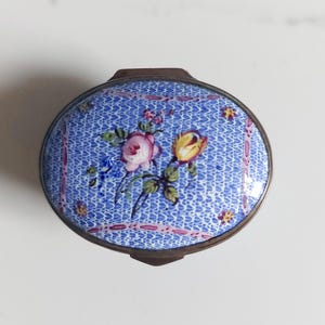 May include: An oval trinket box with a blue patterned lid. The lid is decorated with a floral design in pink and yellow, with green leaves. The box has a metal frame and a blue background.