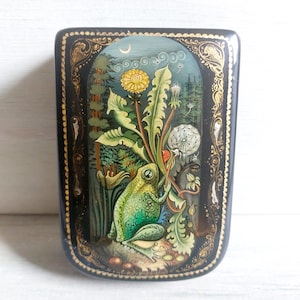 May include: A rectangular, black lacquered box featuring a detailed painting of a green frog amongst dandelions and foliage. The artwork is framed with gold filigree and set against a forest backdrop under a crescent moon.