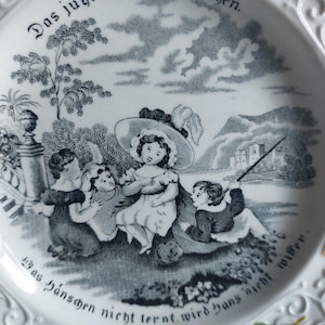 May include: White ceramic plate with a black and white illustration of children playing outdoors. The plate has decorative floral and scrollwork around the rim, and German text. A charming vintage piece.