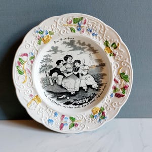 May include: White decorative plate with a scalloped edge and floral accents. The plate features a black and white illustration of children playing, with text above and below the image. The plate has a glossy finish.