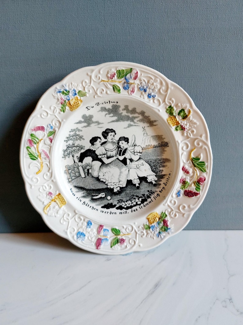May include: White decorative plate with a scalloped edge and floral accents. The plate features a black and white illustration of children playing, with text above and below the image. The plate has a glossy finish.