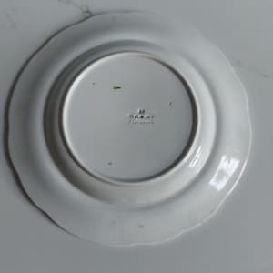 May include: A white ceramic plate with a scalloped edge. The underside of the plate features a central, slightly recessed area with embossed text and markings. The plate's design is simple and elegant, suitable for various dining settings.