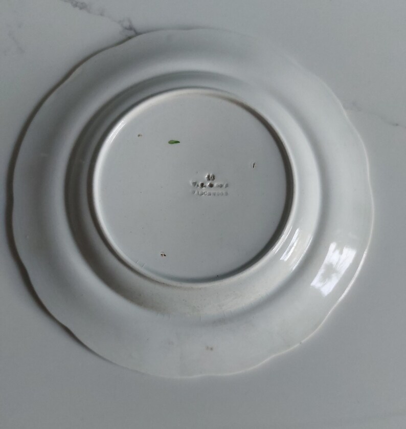 May include: A white ceramic plate with a scalloped edge. The underside of the plate features a central, slightly recessed area with embossed text and markings. The plate's design is simple and elegant, suitable for various dining settings.