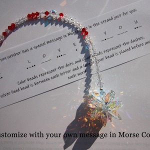May include: A sun catcher with a Morse code message. The strand has clear and red beads, spelling "I LOVE YOU". A clear, multi-faceted flower-shaped crystal hangs below the message. The text reads "Customize with your own message in Morse Code."