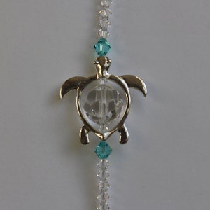 May include: A silver-toned turtle pendant with a clear, faceted center, strung with clear and turquoise crystal beads. The pendant is suspended from a strand of clear beads, creating a decorative ornament.