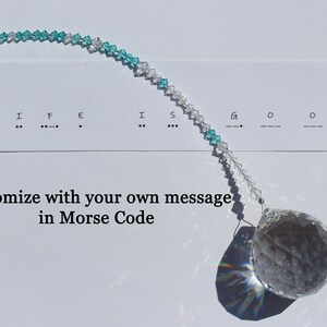 May include: A crystal suncatcher with a clear, faceted sphere and a strand of clear and turquoise beads. The words "LIFE IS GOOD" are written in Morse code. The text "Customize with your own message in Morse Code" is also present.