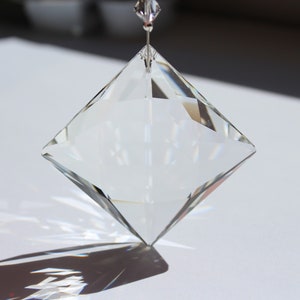 May include: A clear, faceted crystal prism, diamond-shaped, suspended by a thin wire. The crystal reflects light, creating a play of shadows on the white surface below. A small, clear crystal bead is at the top.