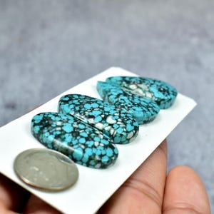 Puede incluir: Cuatro piedras preciosas ovaladas de turquesa con patrones de matriz negros y blancos. Las piedras se muestran en una tarjeta blanca, con una moneda de plata para comparar el tama&ntilde;o. Superficie pulida.
