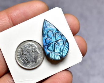 May include: A teardrop-shaped, blue labradorite gemstone with a carved floral design. The gemstone is displayed on a white card next to a U.S. dime coin for size comparison. The gemstone's iridescent quality is visible.