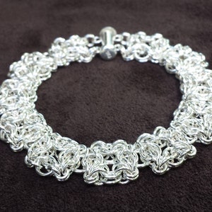 Könnte beinhalten: Eine silberne Kettenkette mit einem filigranen, komplizierten Design. Die Kette besteht aus kleinen, miteinander verbundenen Schlaufen, die ein gewebtes Muster bilden. Die Kette wird mit einem einfachen Verschluss geschlossen.