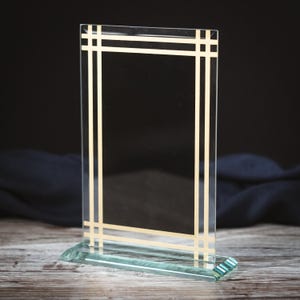 May include: A clear glass plaque with a gold frame. The plaque has a rectangular shape and is standing on a clear glass base.