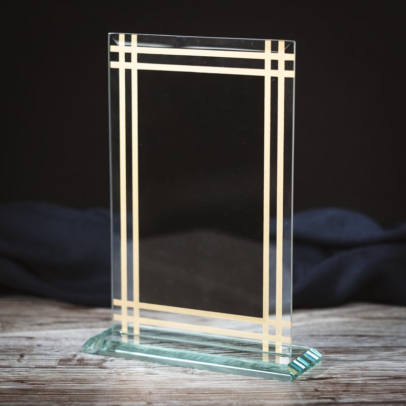 May include: A clear glass plaque with a gold frame. The plaque has a rectangular shape and is standing on a clear glass base.