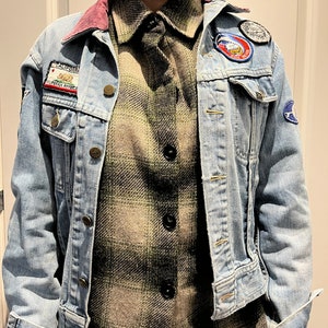 May include: A light blue denim jacket with patches, worn over a green and black plaid button-up shirt. The jacket has a pink collar and brass buttons. Patches include a California bear and a cartoon image.