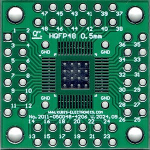 May include: A green circuit board with a black integrated circuit chip in the center. The board has a white label with the text "HQFP48 0.5mm" and the text "GND" printed on it. The board has 48 pins.