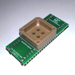 Può includere: Primo piano di un componente elettronico, un adattatore PLCC44T-DIP44, con un socket beige e una scheda a circuito stampato verde. La scheda ha il testo "Q" e "MALIBU-ELECTRONICS.COM". L'adattatore è progettato per dispositivi elettronici.