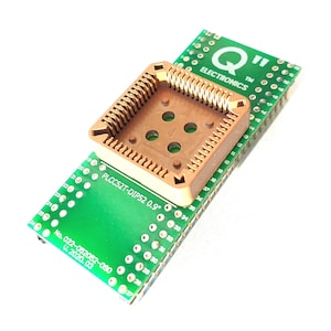 Puede incluir: Una placa de circuito impreso verde con pines dorados y un zócalo de plástico marrón con las inscripciones "PLCC52T-DIP52 0.9"" y "No. 022-052052-090 U. 2020.03". La placa tiene el texto "Q ELECTRONICS TM" impreso en ella.