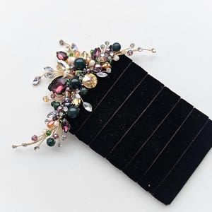 May include: A decorative hair comb featuring an arrangement of dark green, purple, and clear crystals, beads, and rhinestones set on thin gold-toned wire branches. The ornate cluster is attached to a black velvet-covered rectangular hair accessory.