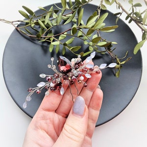 May include: A silver hair comb with a cluster of burgundy and clear crystals, pearls, and beads. The comb is held in a hand against a black background.
