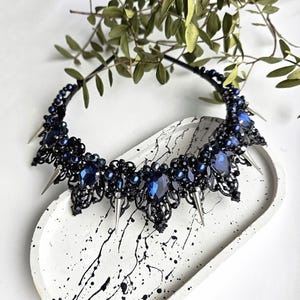 May include: A black headband with a decorative design featuring dark blue gemstones and silver spikes. The ornate piece is embellished with smaller blue beads and is displayed on a white tray with black paint splatters.