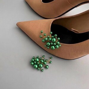 May include: A pair of tan pointed-toe heels with a cluster of green pearl embellishments. The decorative cluster is made of small, round, green pearls attached to thin gold-colored wire, creating a floral-like design. A second cluster is placed near the shoes.