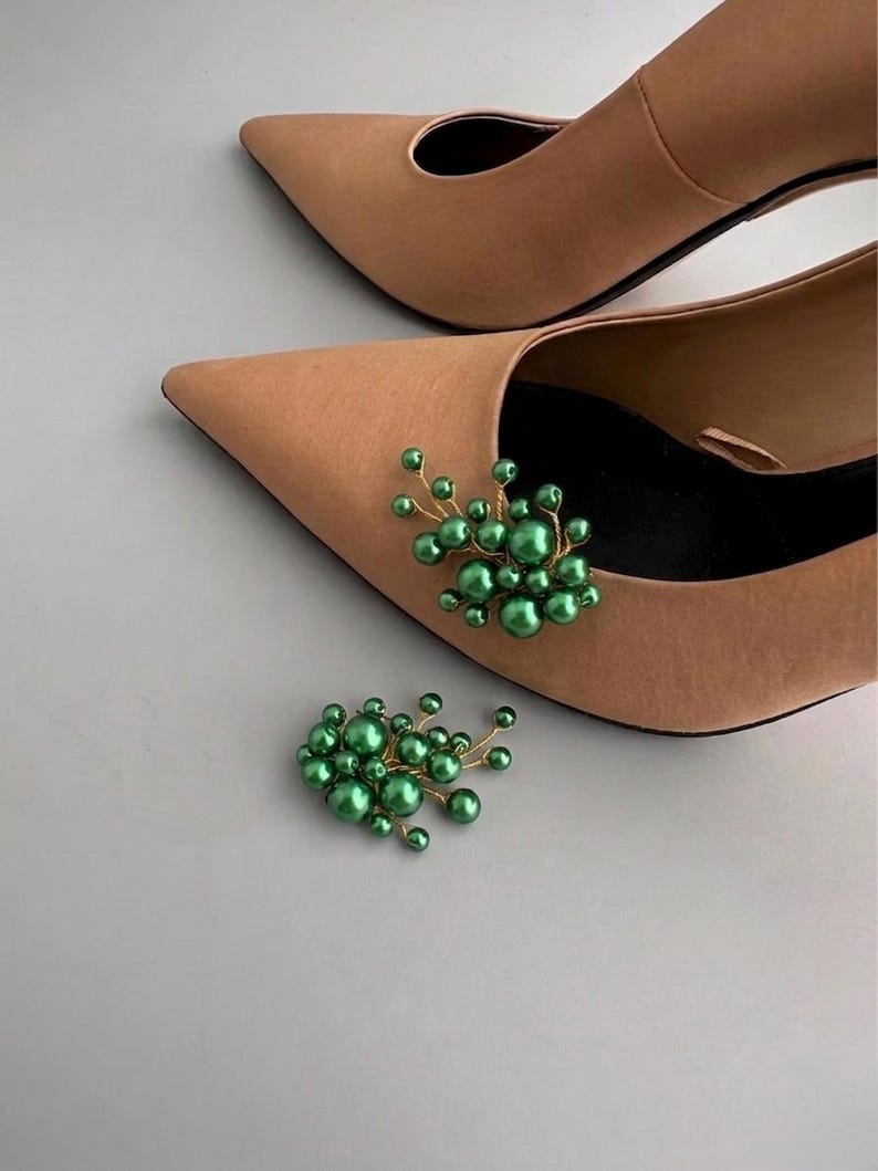 May include: A pair of tan pointed-toe heels with a cluster of green pearl embellishments. The decorative cluster is made of small, round, green pearls attached to thin gold-colored wire, creating a floral-like design. A second cluster is placed near the shoes.