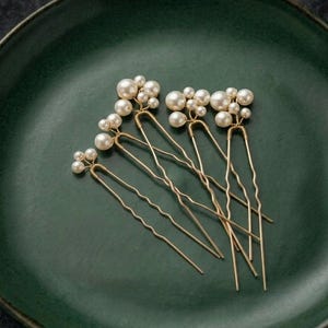 May include: A collection of six gold-toned hair pins, each adorned with clusters of faux pearls in varying sizes. The pins are arranged on a dark green ceramic plate, creating a contrast between the delicate accessories and the rustic background.