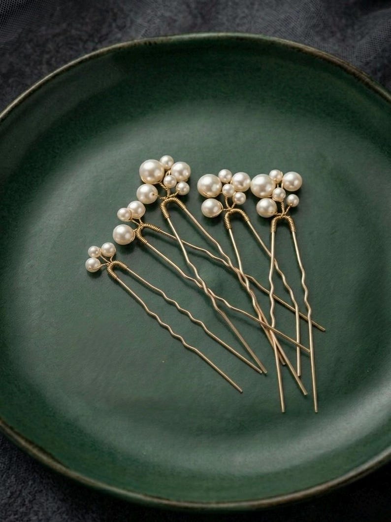 May include: A collection of six gold-toned hair pins, each adorned with clusters of faux pearls in varying sizes. The pins are arranged on a dark green ceramic plate, creating a contrast between the delicate accessories and the rustic background.