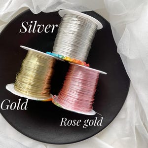 May include: Three spools of wire in silver, gold, and rose gold are stacked on a black plate. The word "Silver" is written above the silver spool, "Gold" is written near the gold spool, and "Rose gold" is written near the rose gold spool. White fabric is in the background.