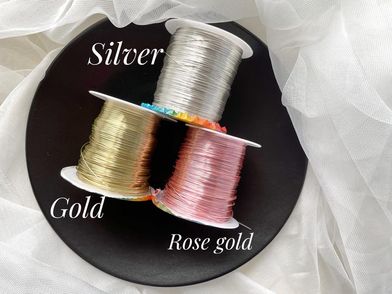 May include: Three spools of wire in silver, gold, and rose gold are stacked on a black plate. The word "Silver" is written above the silver spool, "Gold" is written near the gold spool, and "Rose gold" is written near the rose gold spool. White fabric is in the background.