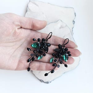 May include: A pair of dangle earrings featuring black wire branches adorned with small black beads and faceted emerald green teardrop crystals. The earrings have a hook-style closure and a cluster design that extends downward in a delicate, organic shape.