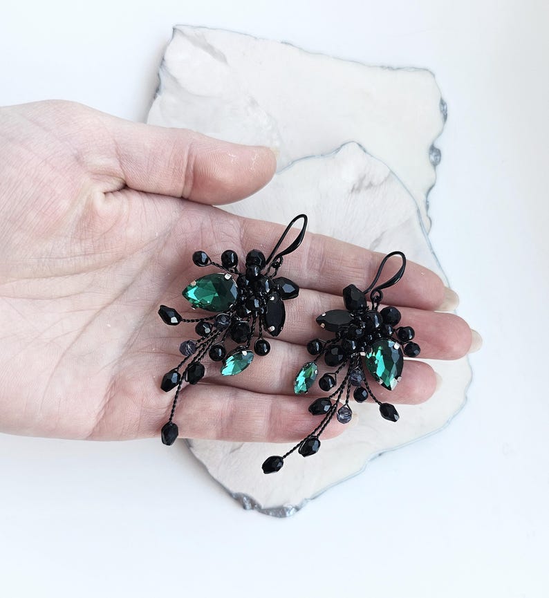 May include: A pair of dangle earrings featuring black wire branches adorned with small black beads and faceted emerald green teardrop crystals. The earrings have a hook-style closure and a cluster design that extends downward in a delicate, organic shape.