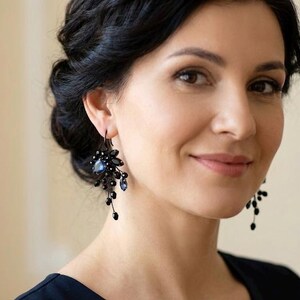 May include: A pair of dangling earrings featuring black metal wire, small black beads, and a central dark blue teardrop-shaped crystal. The earrings have a cluster design with multiple thin strands extending downward, creating a layered appearance.